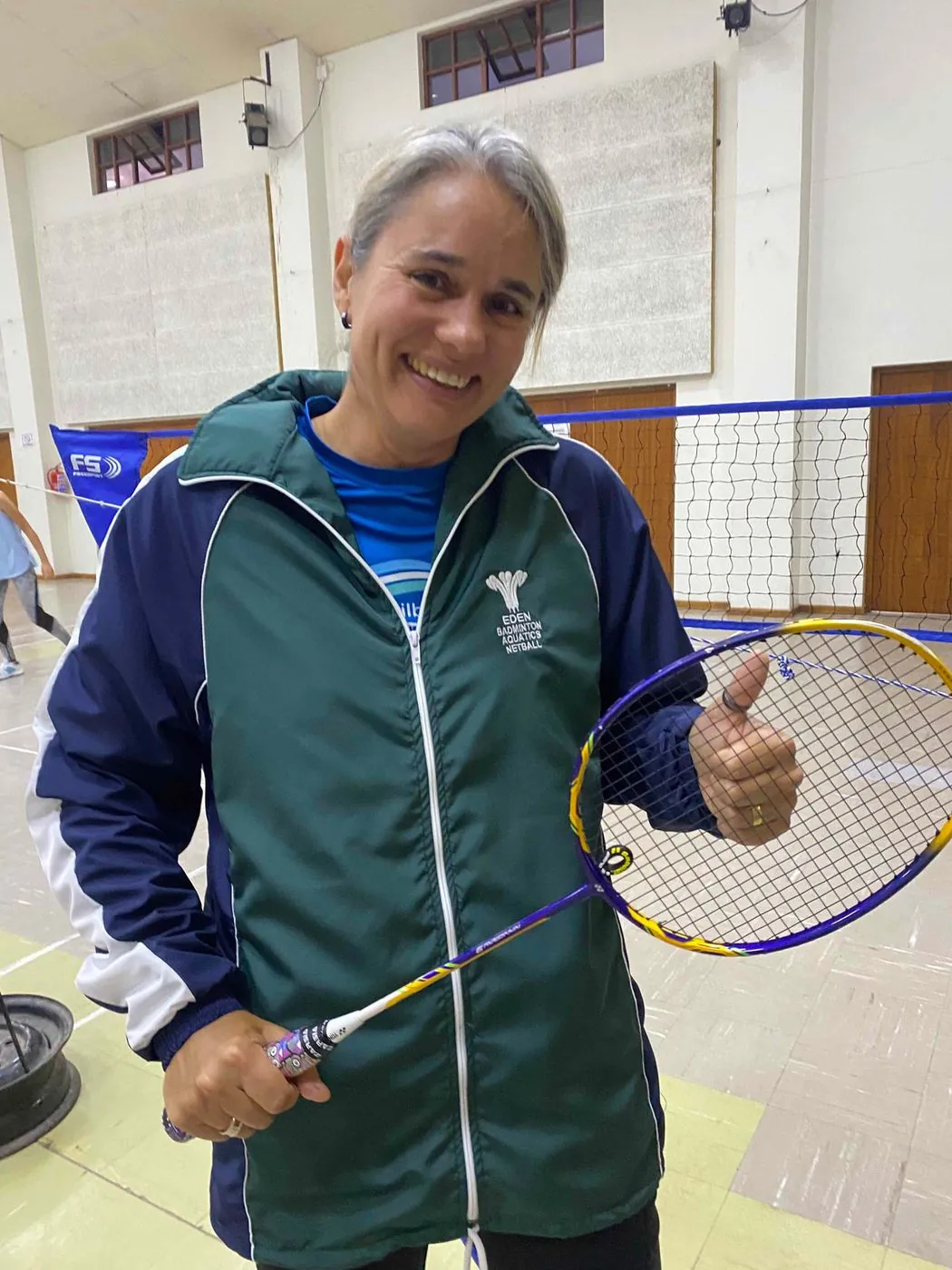 A smiling woman in a green tracksuit jacket holds a badminton racket and gives a thumbs up in a hall.
