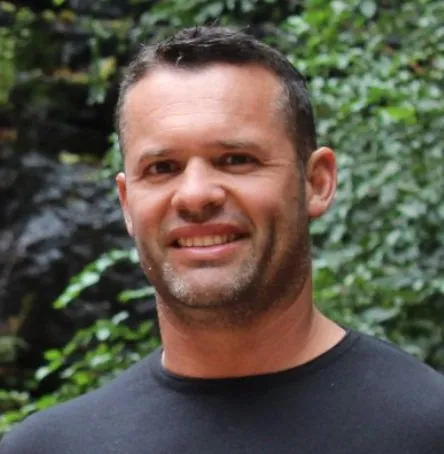A smiling man in a dark T-shirt in front of green foliage.
