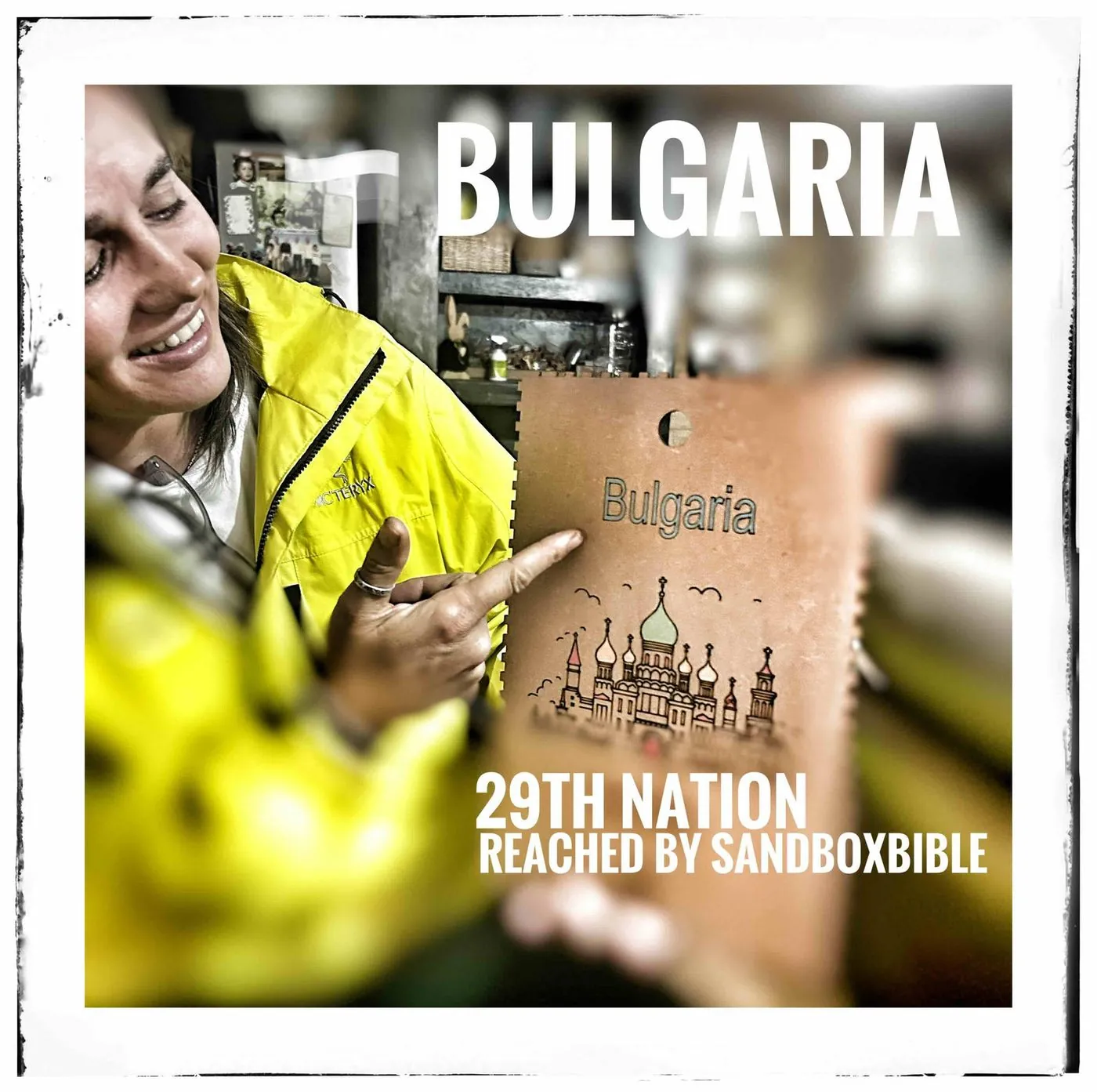 A woman in a yellow jacket holds an engraved wooden tag reading Bulgaria, with the text Bulgaria, 29th nation reached by SandBoxBible.
