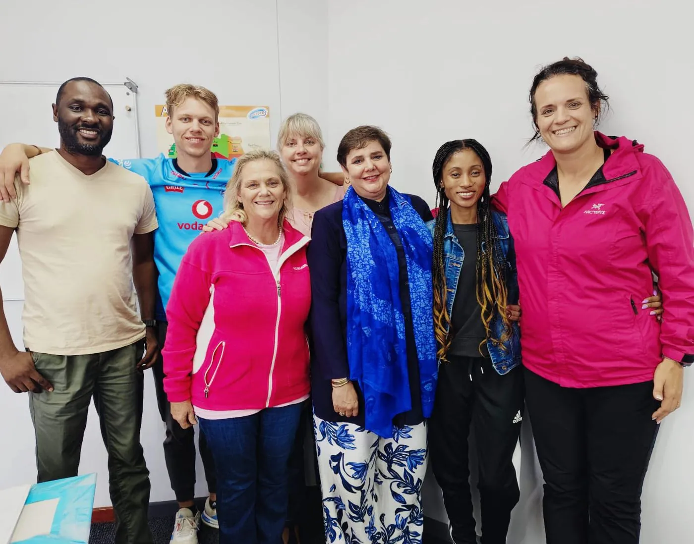 Eight women and two men standing together in a bright room, several in pink jackets.