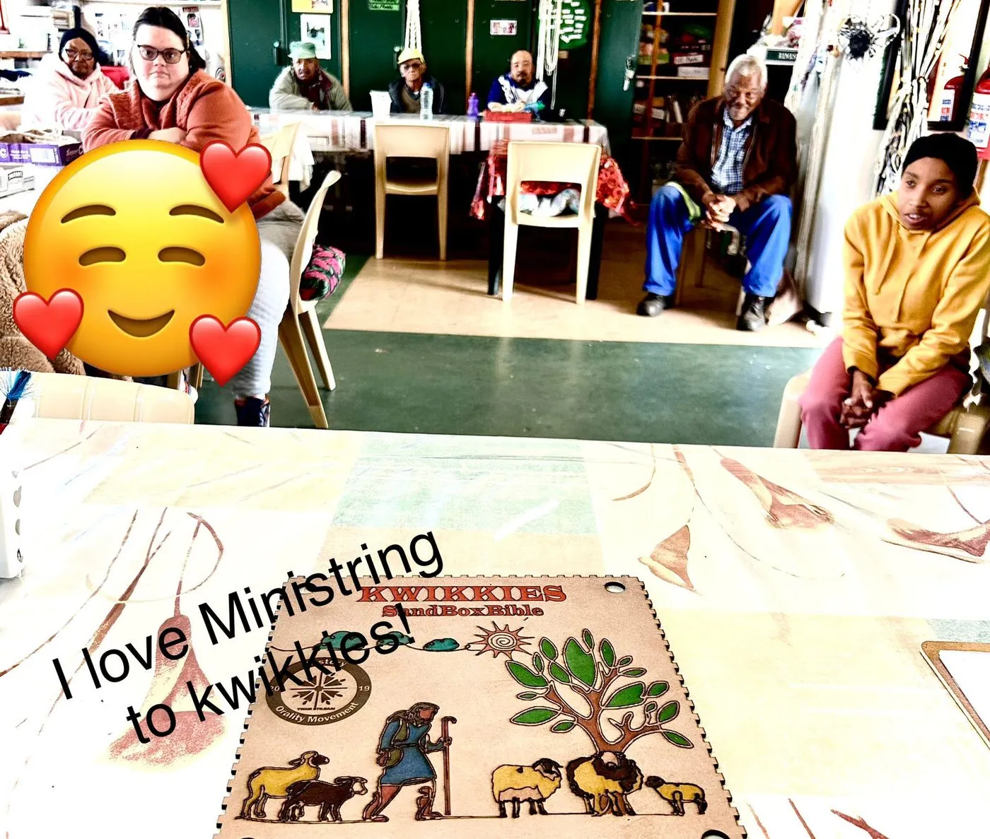 A dining area with people at tables and a painted wooden SandBoxBible board, captioned I love Ministring to kwikkies.
