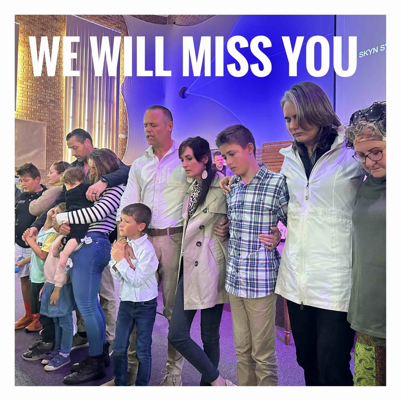 A family of adults and children standing with their arms around each other on a stage, with the words We will miss you.