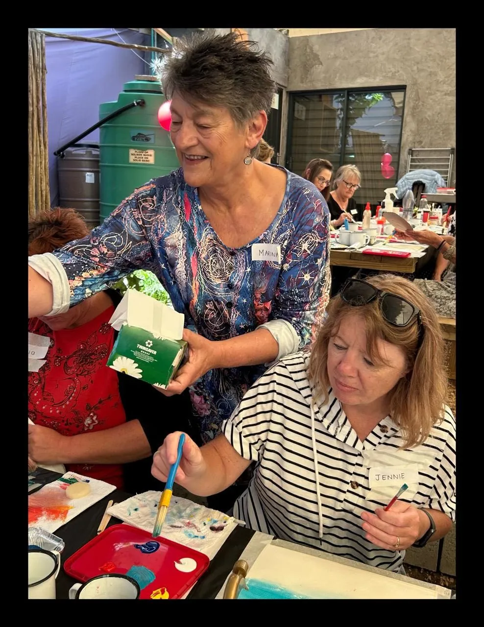 Several women sitting and standing around a table, painting and handling art supplies.