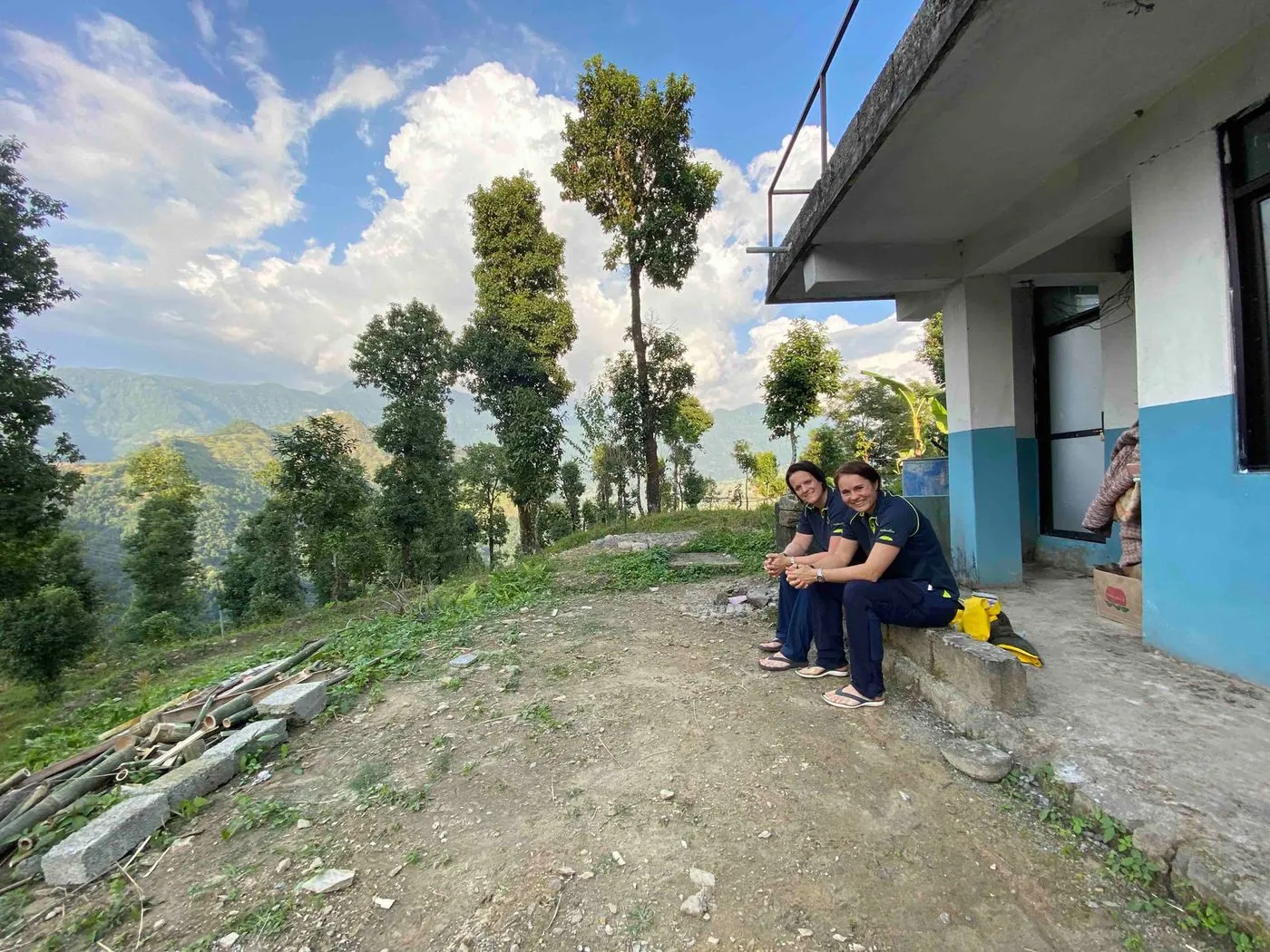 Two people sitting on the edge of a dirt path beside a building, with a green valley and trees behind.