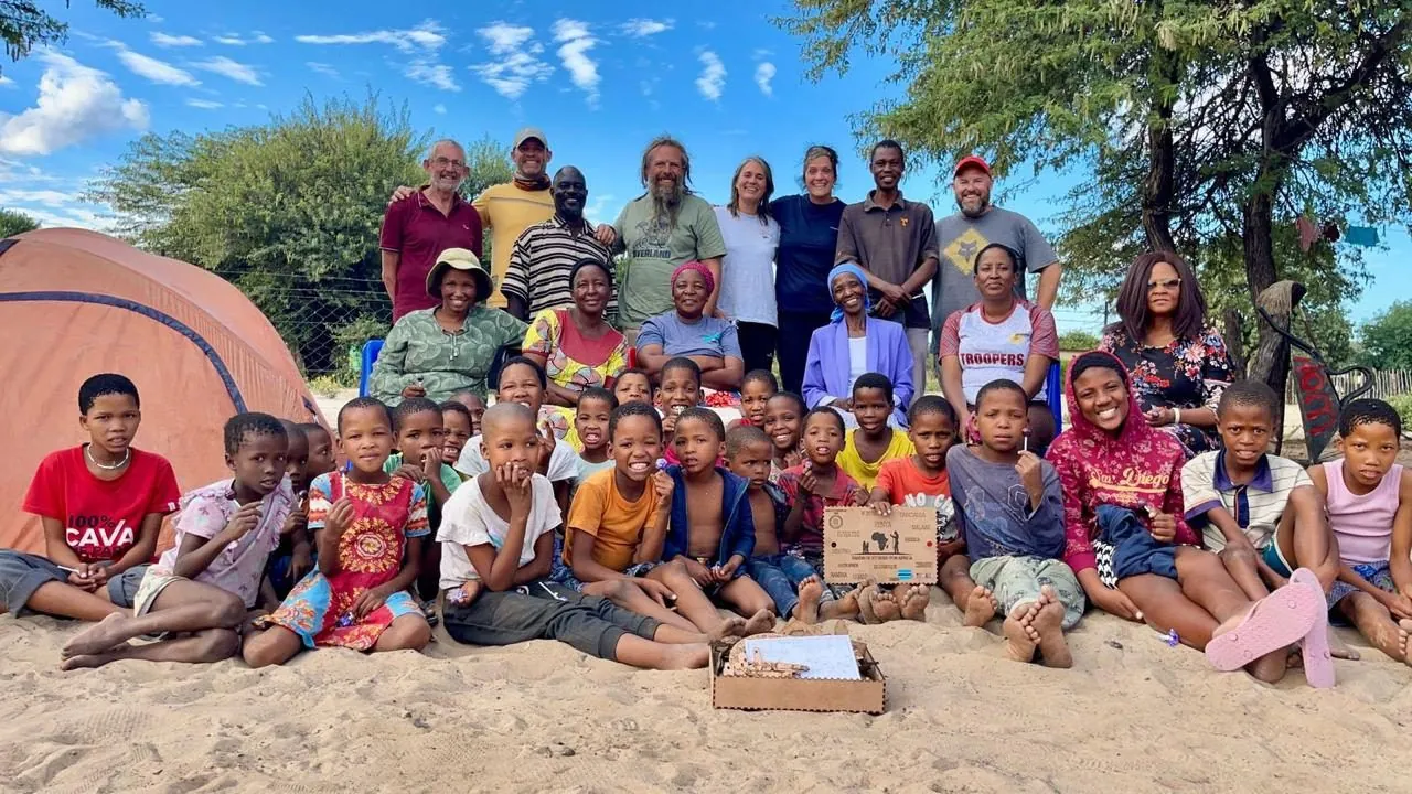 A large group of adults and children sitting and standing together on sand, with a box in front of them.