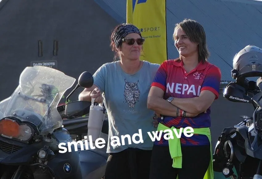 Two smiling women in riding gear beside motorbikes, captioned Smile and wave.