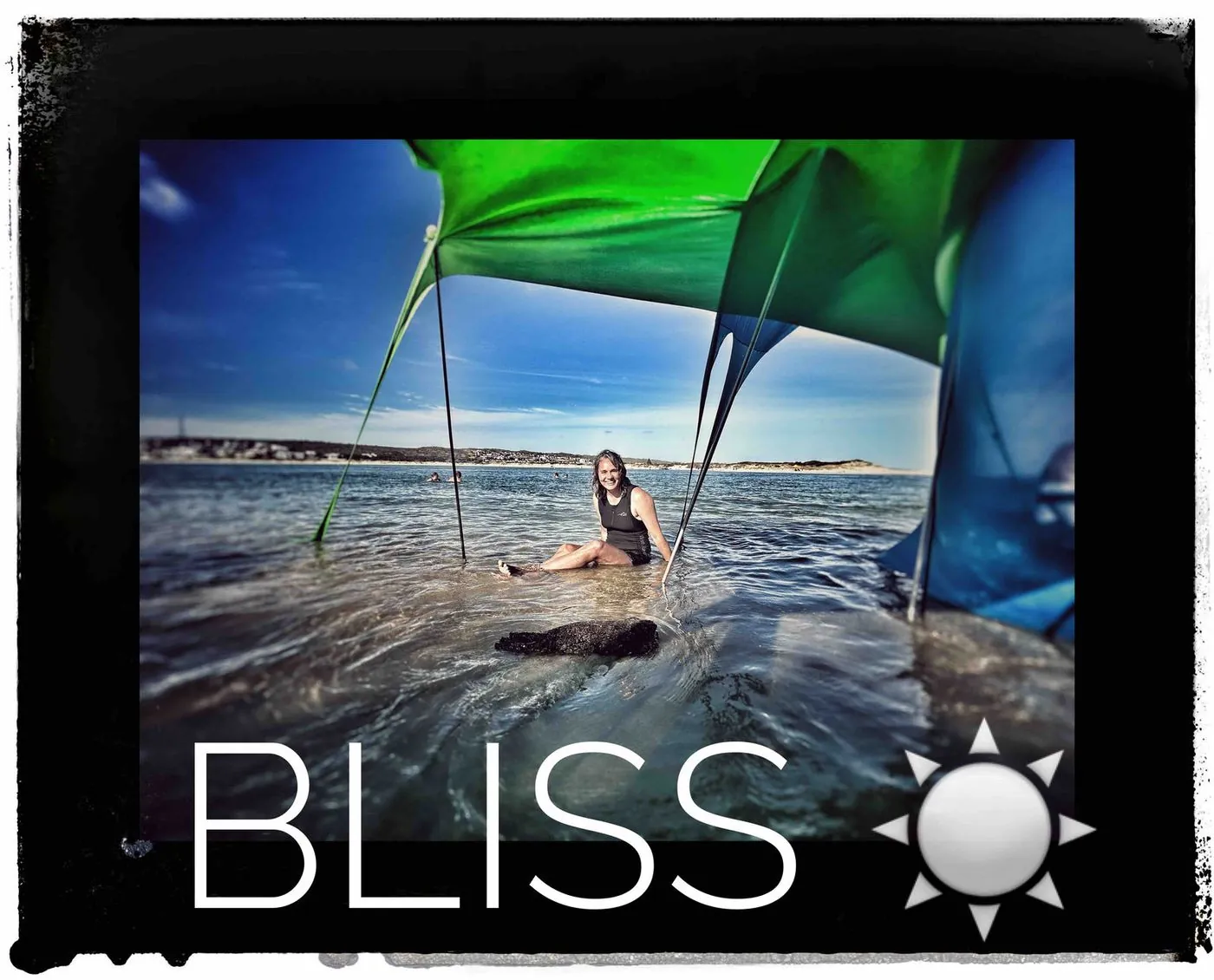 A woman sitting in shallow water under a blue tarp shelter, captioned Bliss.