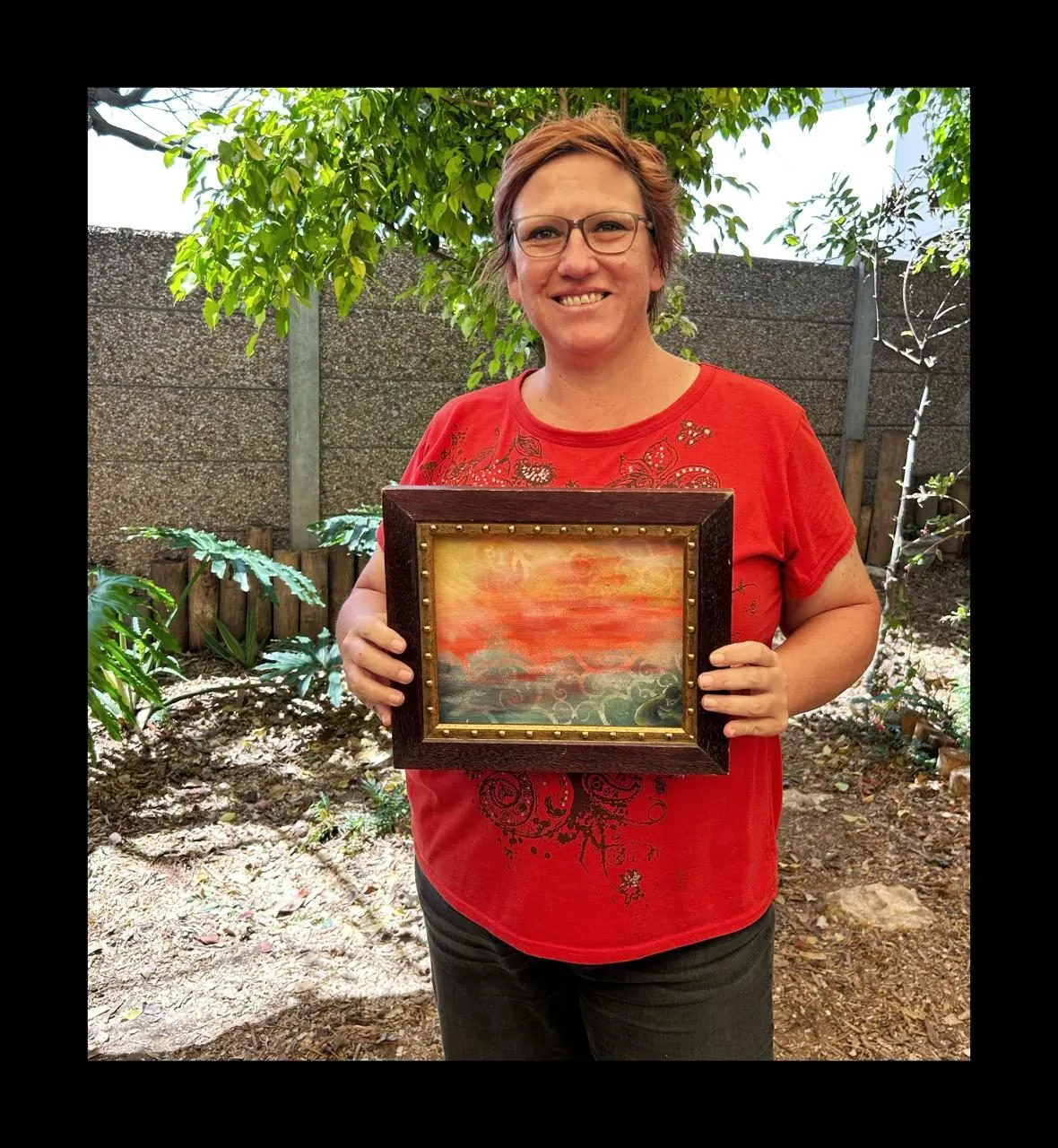 A smiling woman in a red top holds a framed painting of a red-orange sunset, standing in a garden.
