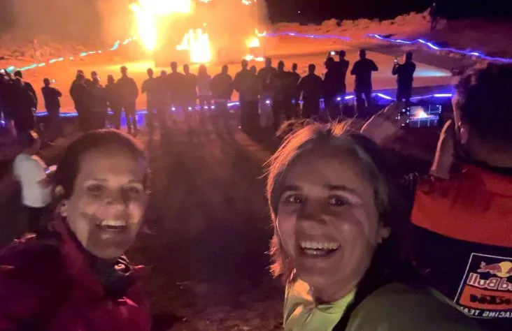 Two women taking a selfie at night with a large fire and a crowd behind them.