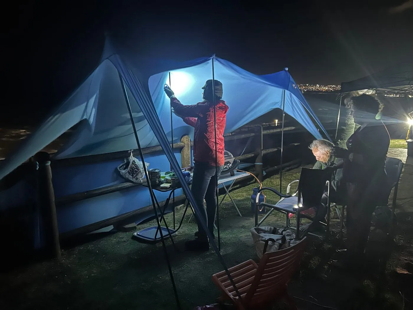 A blue tarp shelter lit at dusk with a person sitting beneath it.