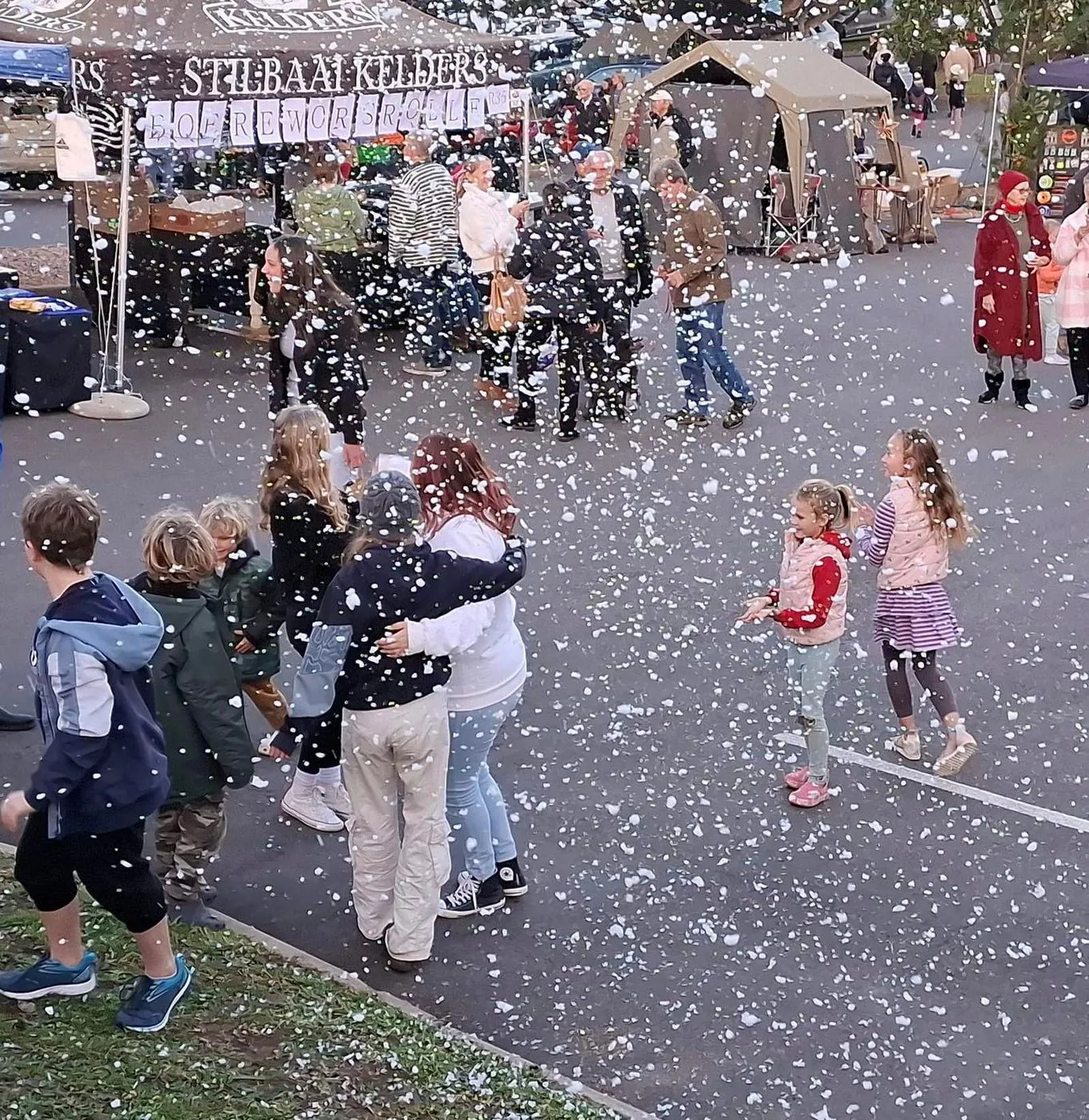 A crowd of adults and children in a street under falling white confetti, with market tents and a banner behind them.