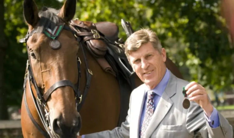 A man in a light suit stands beside a bridled brown horse, holding a medal.