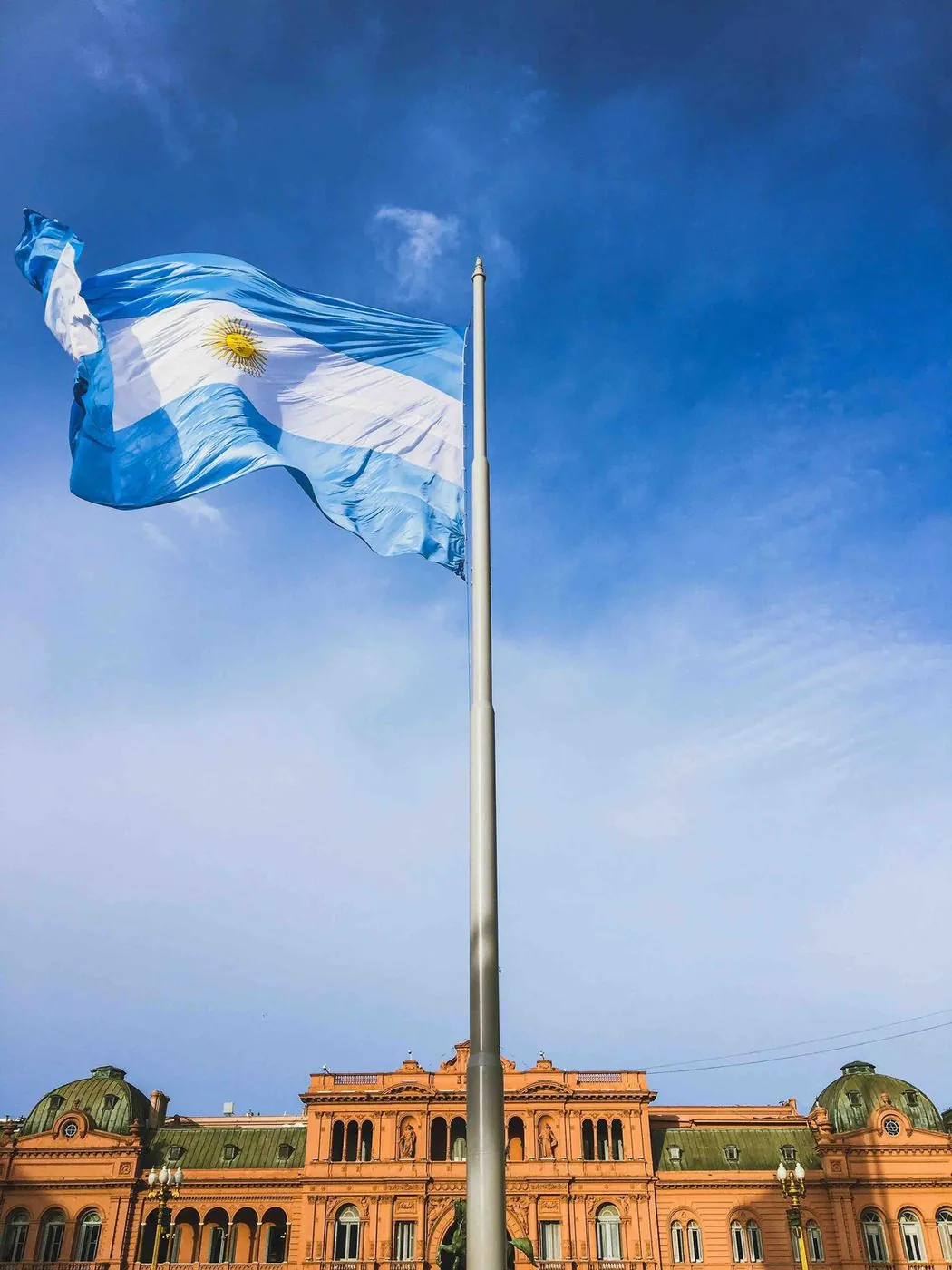 An Argentine flag on a pole in front of a pink government building.