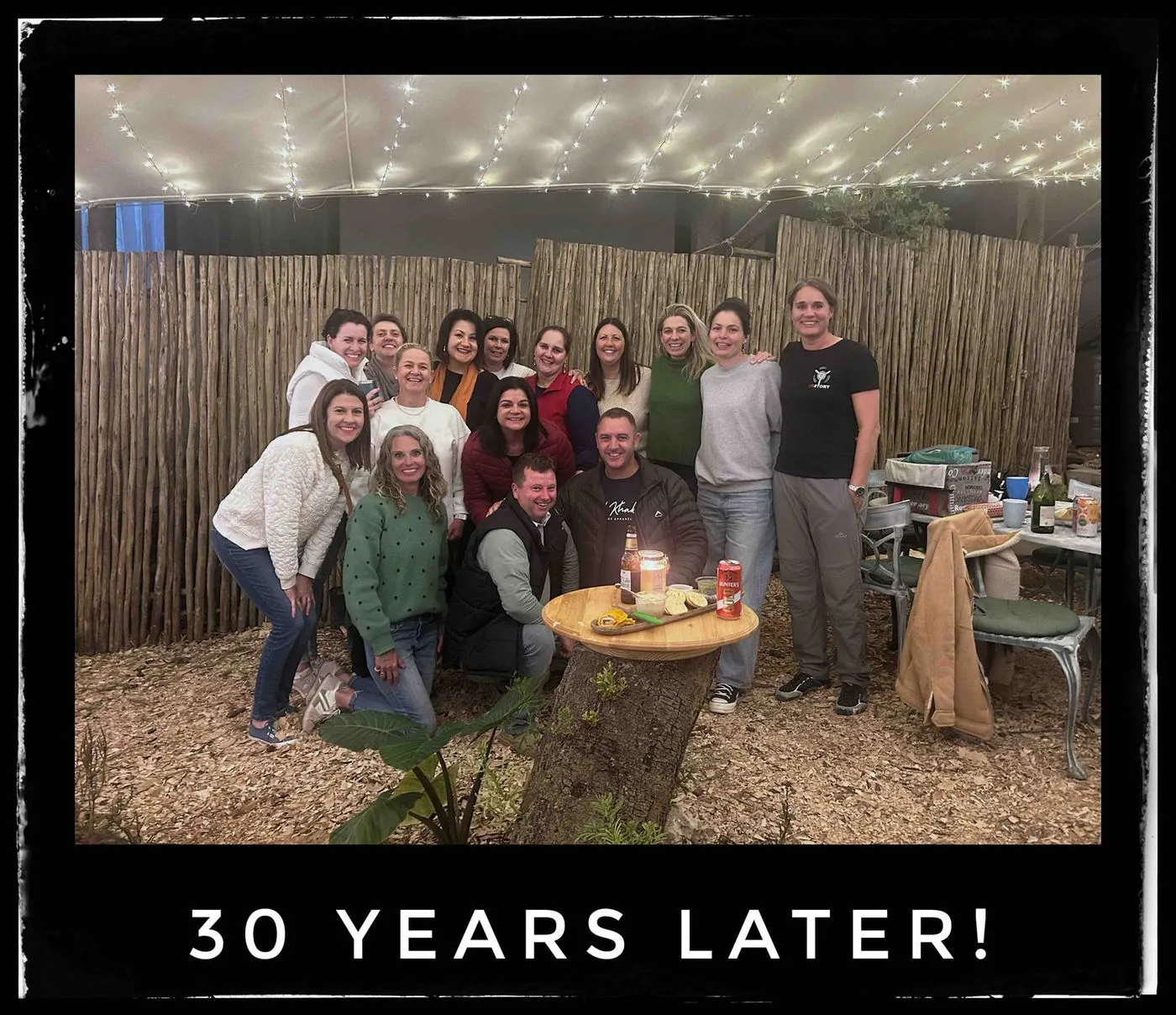 About a dozen women and men posing together in front of a reed screen and a table, captioned 30 years later!