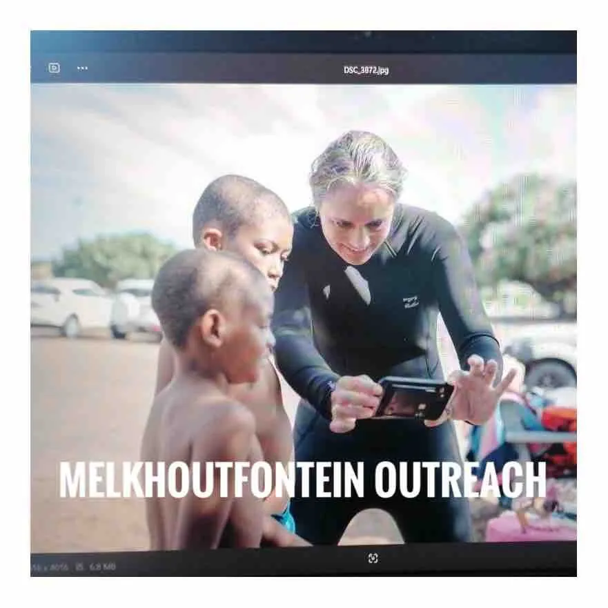 A woman in a wetsuit crouches with two young boys looking at a phone, captioned Melkhoutfontein outreach.