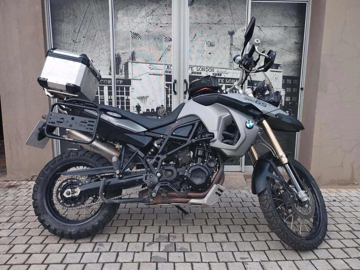 A silver and black BMW adventure motorbike with a top box parked on paving in front of glass doors.