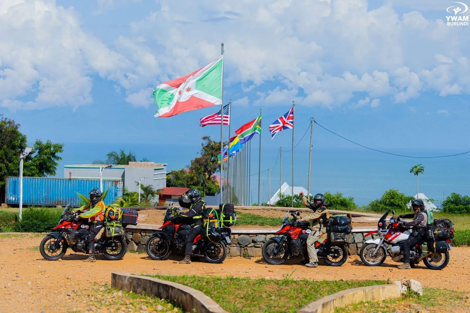 Four riders on loaded motorbikes parked in a row beside a low stone wall, with a line of national flags on tall poles, the sea and a blue sky behind them.