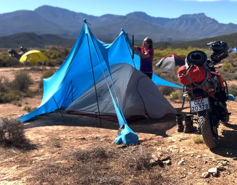 A blue tarp shelter pitched beside a small grey tent on dry scrubland, with a person standing at its pole and a loaded red motorbike parked next to it, and a mountain range behind.