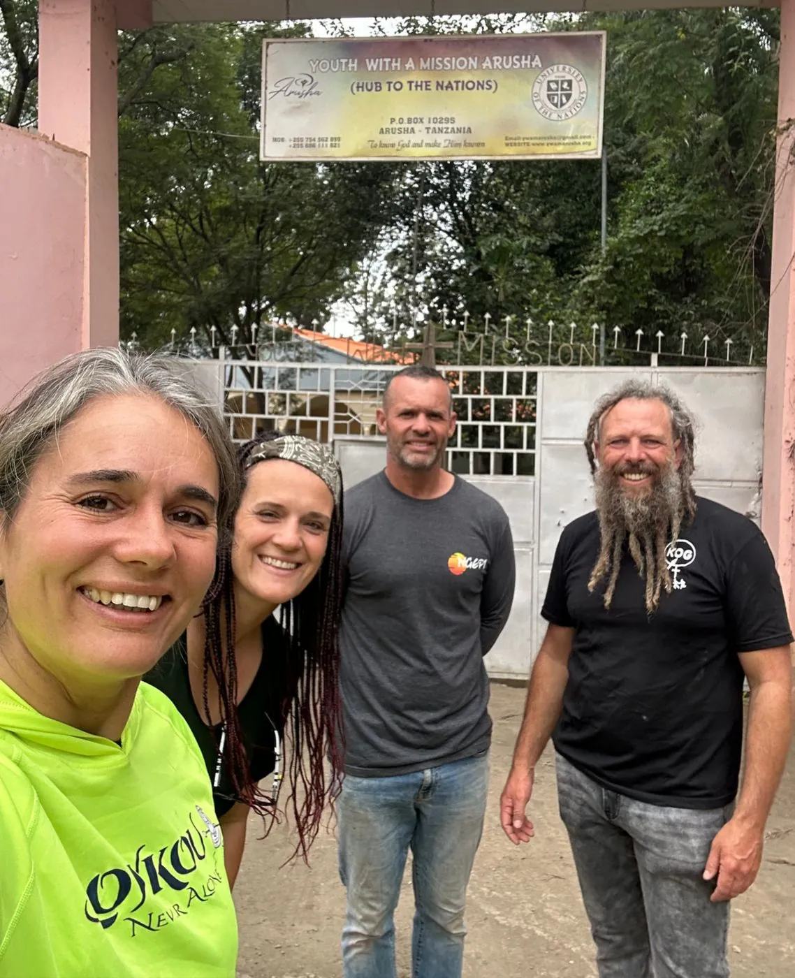 Four people standing together and smiling in front of a gate with a sign; one wears a bright yellow-green shirt.