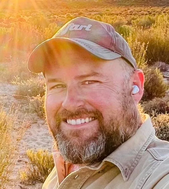 A smiling man in a cap and khaki shirt taking a selfie at sunset in dry grass.