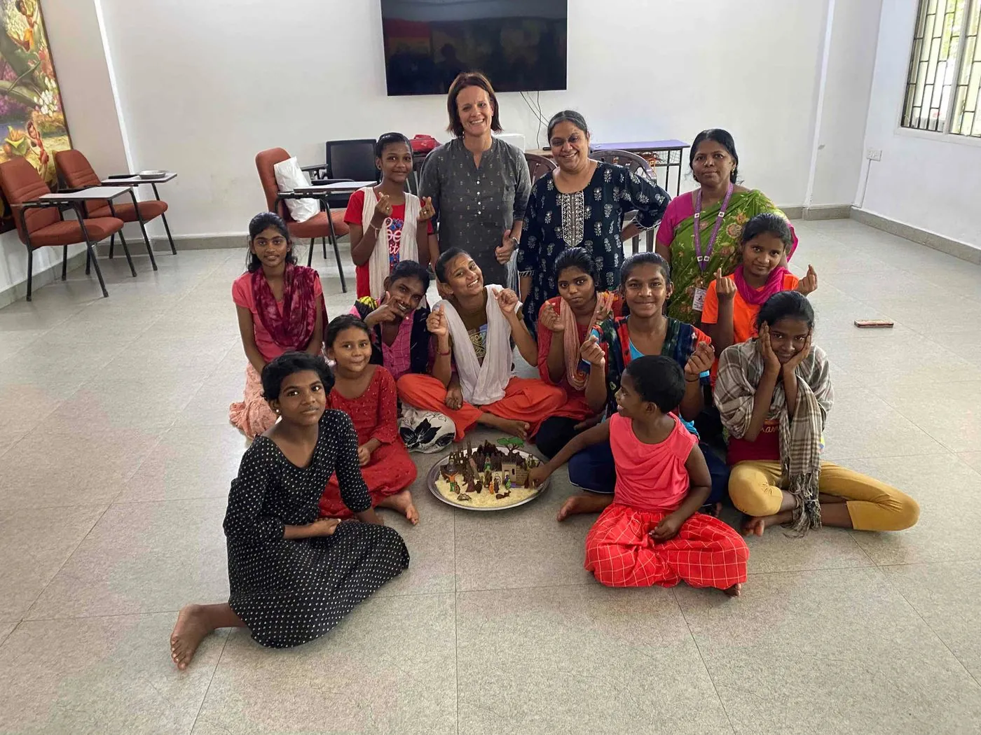 A group of girls and women sitting on the floor of a room around a low tray, smiling at the camera.