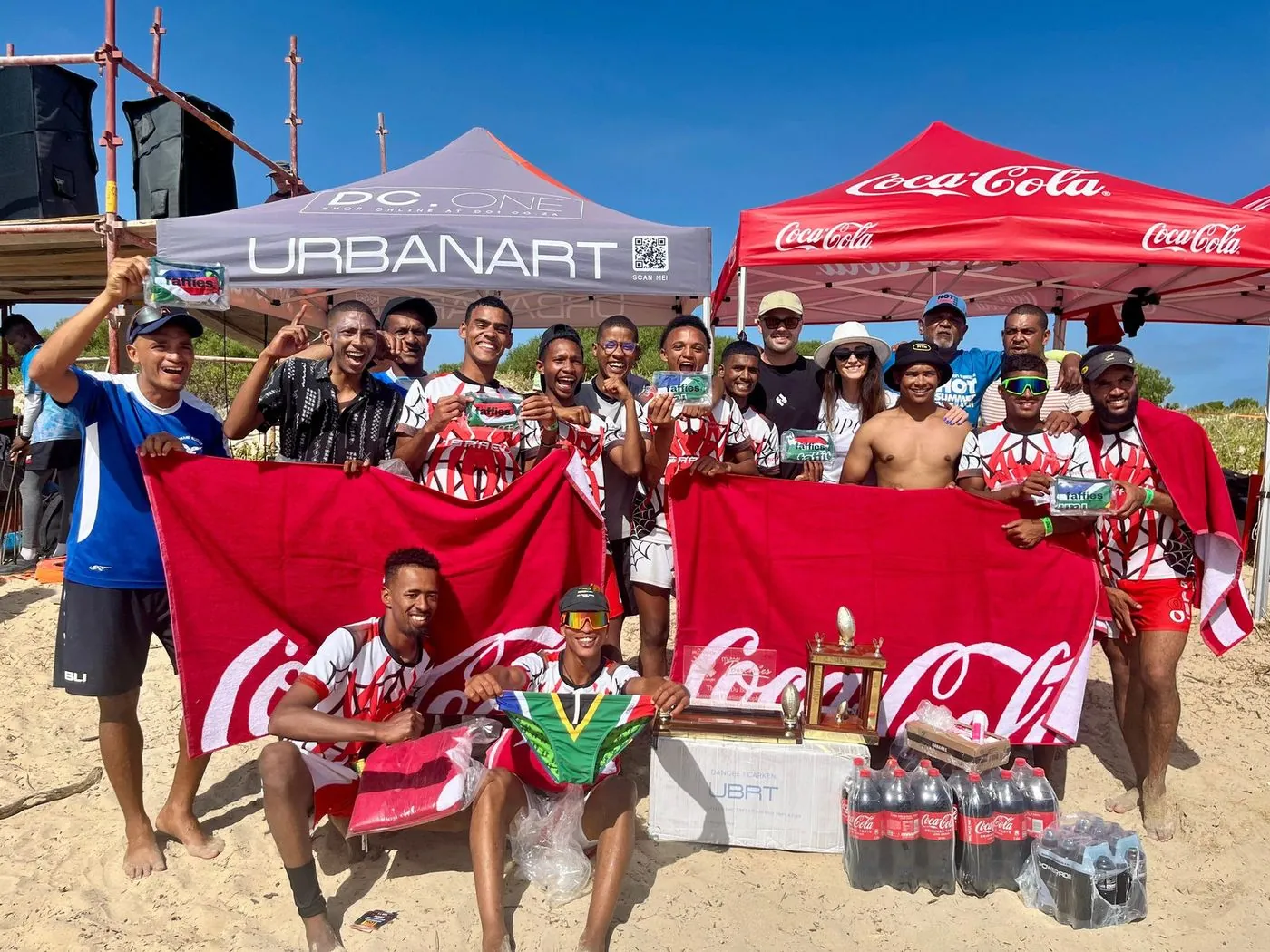 A crowd of players and spectators holding red flags in front of a Coca-Cola beach tent.