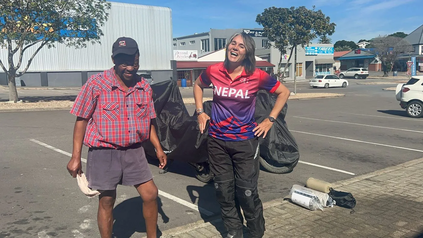 A man in a checked shirt and a woman in a red Nepal top stand on a street next to a motorbike.