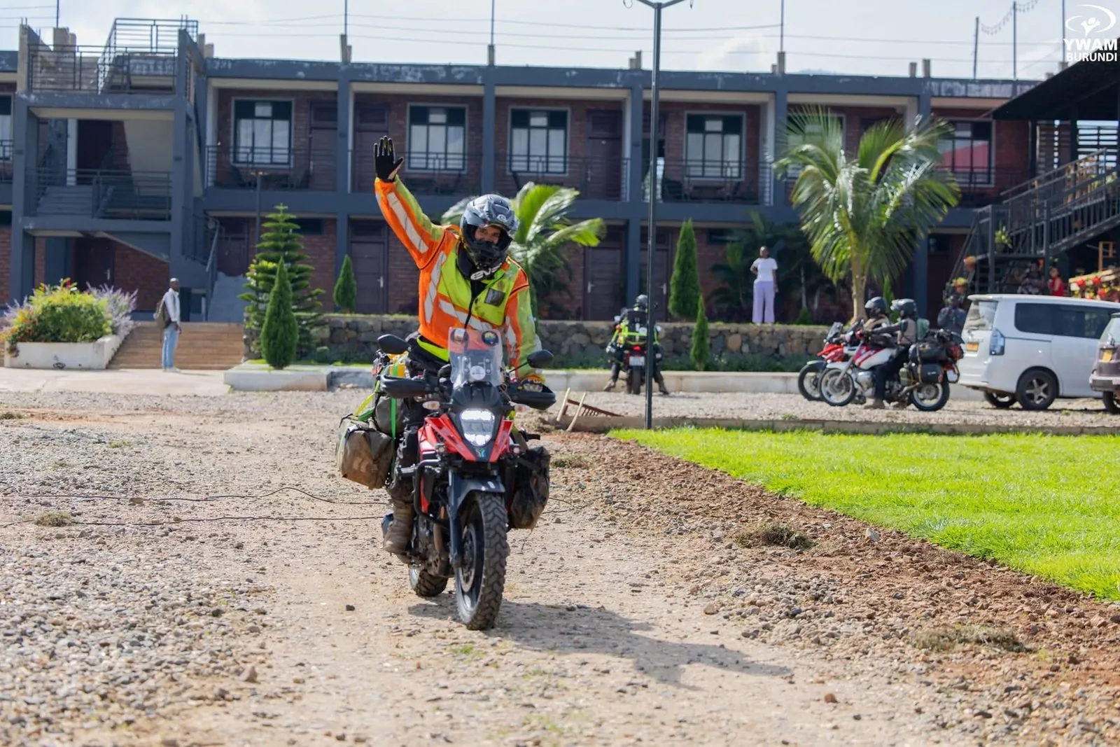 A rider in an orange and yellow high-visibility vest and a grey helmet raises one gloved hand while riding a loaded red and black adventure motorbike along a gravel path, with a two-storey brick building, palm trees and other parked motorbikes behind.