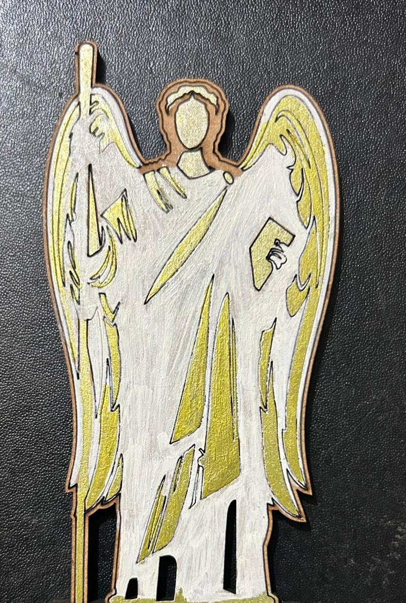 A flat wooden figure of an angel with white and gold-painted wings and robe, holding a staff, photographed on a dark surface.