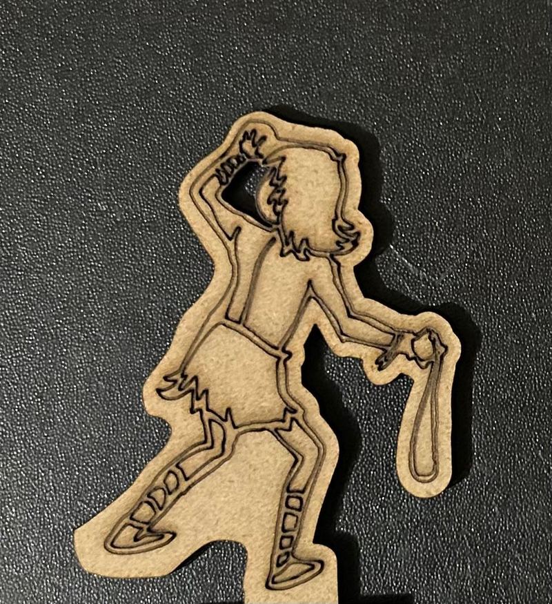 A flat laser-cut wooden figure of a young man in a short tunic holding a sling in one hand with the other hand raised to his head, photographed on a dark surface.