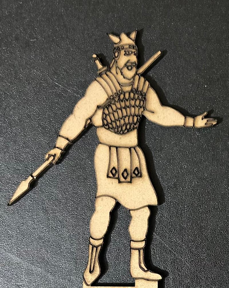 A flat laser-cut wooden figure of a bearded warrior in scale armour and a horned helmet, holding a spear and reaching out one hand, photographed on a dark surface.