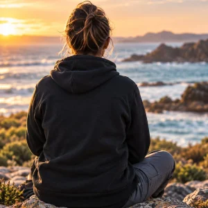 A person seen from behind, sitting on lichen-covered rocks in a black hoodie and looking out at the sea and a sunset sky.