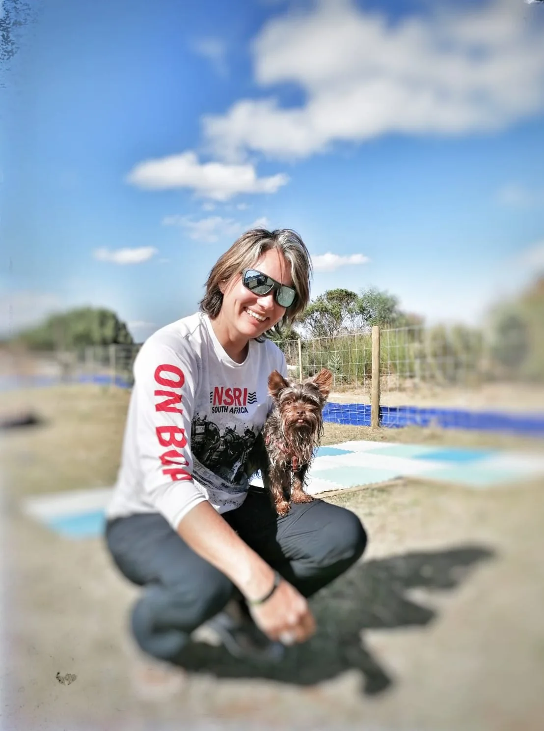 Sarah smiles at the camera in sunglasses and a white long-sleeved shirt, crouching outdoors with a small brown and tan dog beside her, with a blue sky and scattered clouds behind.