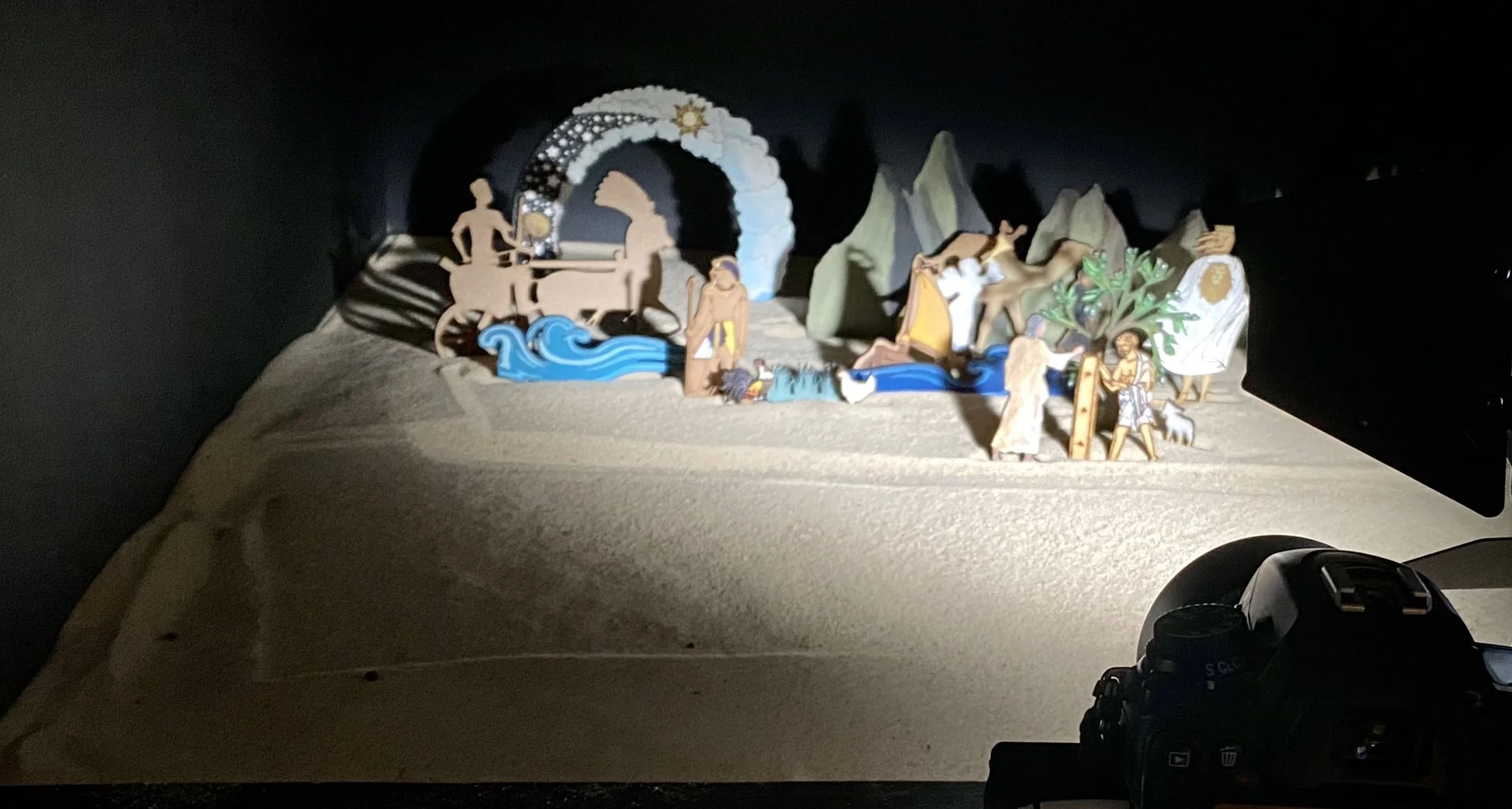 A small set of flat painted figures, blue waves, green rocks and a tree standing in pale sand, lit by a studio light in a dark room, with the top of a camera in the bottom corner.