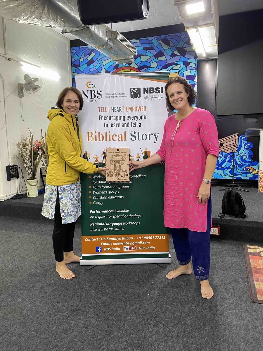 Two women stand side by side in a hall with a colourful mural, holding a wooden SandBoxBible plaque between them in front of a tall banner that reads Tell, Hear, Empower: Encouraging everyone to learn and tell a Biblical Story.
